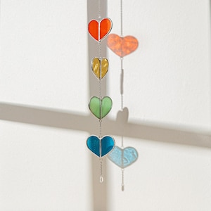 Op de afbeelding: Een hangende decoratie met vier hartvormige glas-in-lood panelen in oranje, geel, groen en blauw. De harten zijn verbonden door een zilveren ketting. Een kleurrijk, opvallend ornament.