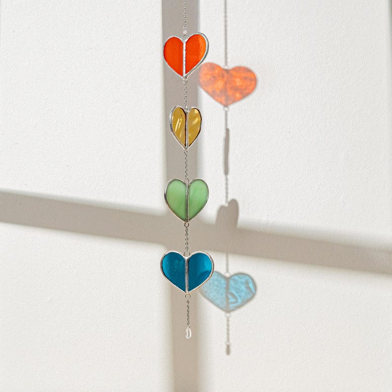 Op de afbeelding: Een hangende decoratie met vier hartvormige glas-in-lood panelen in oranje, geel, groen en blauw. De harten zijn verbonden door een zilveren ketting. Een kleurrijk, opvallend ornament.