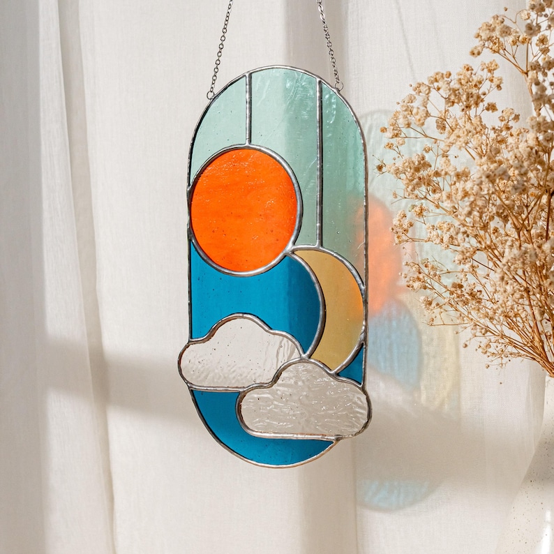 K&ouml;nnte beinhalten: Ein Buntglasfenster mit einem Sonnen-, Mond- und Wolkenmotiv in einem Farbschema aus T&uuml;rkis, Orange und Gelb. Das Buntglas ist oval geformt und h&auml;ngt an einer silbernen Kette.