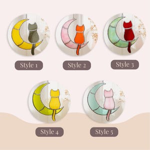 May include: Five different stained glass cat moon ornaments. Each ornament features a cat sitting on a crescent moon. The cats are in different colors: black, orange, red, yellow, and pink. The moons are also in different colors: yellow, pink, green, and blue.