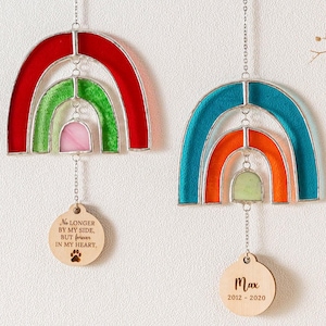 May include: Two rainbow stained glass suncatchers with wooden tags hanging from them. The top suncatcher is red, green, and pink. The bottom suncatcher is blue, orange, and green. The wooden tags have the text "No longer by my side, but forever in my heart." and "Max 2012-2020".
