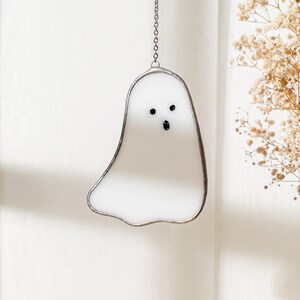 May include: A white stained glass ghost with black eyes and mouth, hanging from a silver chain. The ghost is outlined in silver and has a simple, friendly design. Perfect for Halloween or spooky decor.