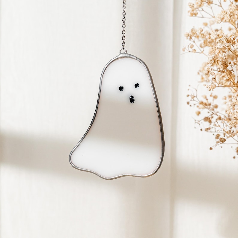May include: A white stained glass ghost with black eyes and mouth, hanging from a silver chain. The ghost is outlined in silver and has a simple, friendly design. Perfect for Halloween or spooky decor.
