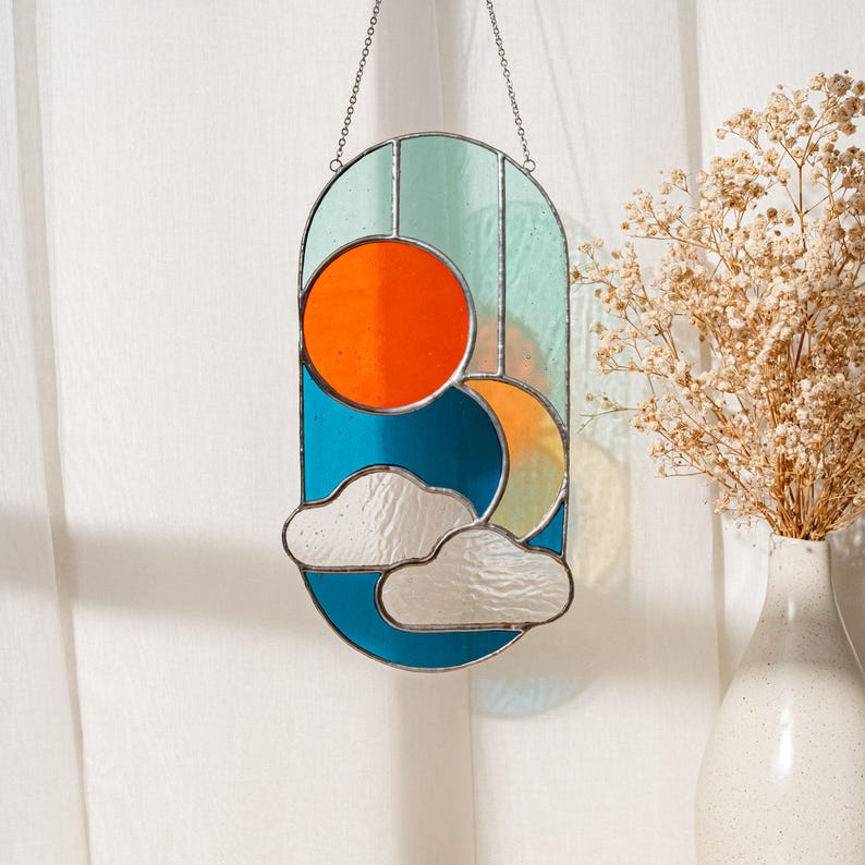 K&ouml;nnte beinhalten: Ein Buntglasfenster mit Sonne und Mond, das an einer silbernen Kette h&auml;ngt. Die Sonne ist orange, der Mond ist gelb und die Wolken sind wei&szlig; vor einem blauen und gr&uuml;nen Hintergrund.