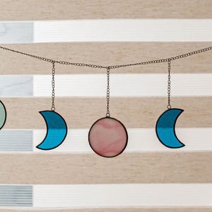Puede incluir: Una guirnalda decorativa de fases lunares con lunas crecientes y una luna llena. Las lunas son de color blanco, azul y verde, con una luna llena rosa en el centro. Las lunas est&aacute;n suspendidas de una cadena.