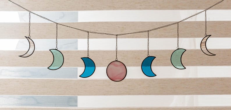 Puede incluir: Una guirnalda decorativa de fases lunares con lunas crecientes y una luna llena. Las lunas son de color blanco, azul y verde, con una luna llena rosa en el centro. Las lunas est&aacute;n suspendidas de una cadena.