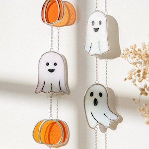 May include: Halloween-themed hanging decorations featuring orange pumpkins and white ghosts with smiling faces. The decorations are made of stained glass and are suspended by silver chains. The pumpkins and ghosts are in a vertical arrangement.