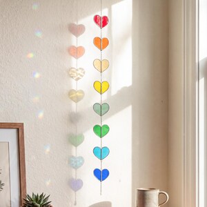 Op de afbeelding: Een hangende hartjesslinger van glas-in-lood in regenboogkleuren. De harten zijn opgehangen aan een zilveren ketting, met kleuren als rood, oranje, geel, groen en blauw. De slinger hangt bij een raam, met kleurrijke lichtreflecties.