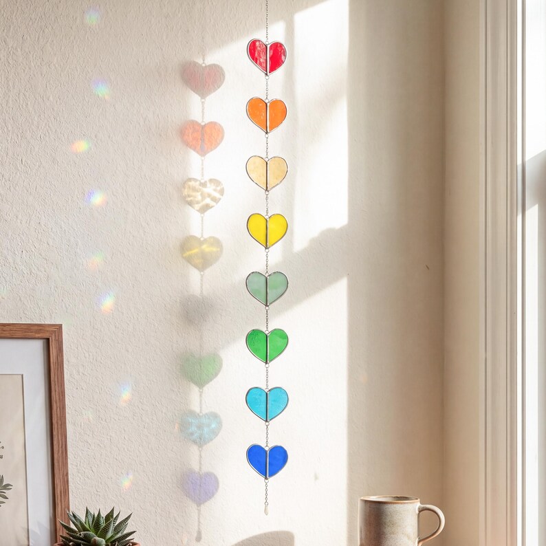 Op de afbeelding: Een hangende hartjesslinger van glas-in-lood in regenboogkleuren. De harten zijn opgehangen aan een zilveren ketting, met kleuren als rood, oranje, geel, groen en blauw. De slinger hangt bij een raam, met kleurrijke lichtreflecties.