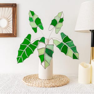 May include: A stained glass plant arrangement featuring five green leaves in varying shades, set in a cream-colored vase. The leaves are arranged in a fan shape, with a woven placemat beneath the vase. The leaves are made of glass and are outlined with a silver-colored metal.