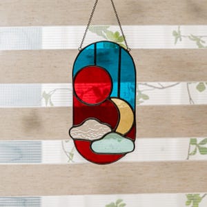 K&ouml;nnte beinhalten: Ein Buntglas-Sonnenf&auml;nger mit einem roten, gelben, blauen und wei&szlig;en Design. Der Sonnenf&auml;nger zeigt eine rote Sonne, einen gelben Halbmond und wei&szlig;e Wolken vor blauem Hintergrund.