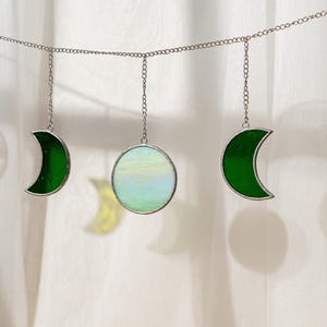 Puede incluir: Una guirnalda decorativa con adornos de fases lunares de vidrio. La guirnalda incluye medias lunas verdes y amarillas, y una luna llena en azul p&aacute;lido y blanco. Las piezas de vidrio est&aacute;n enmarcadas en plata y suspendidas por cadenas.