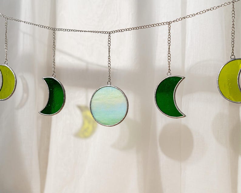 Puede incluir: Una guirnalda decorativa con adornos de fases lunares de vidrio. La guirnalda incluye medias lunas verdes y amarillas, y una luna llena en azul p&aacute;lido y blanco. Las piezas de vidrio est&aacute;n enmarcadas en plata y suspendidas por cadenas.