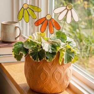 Op de afbeelding: Een terracotta keramische pot met een decoratief patroon bevat een groen-witte plant. Drie kleurrijke glas-in-lood bloemstokken in oranje, wit en groen zijn in de plant gestoken.