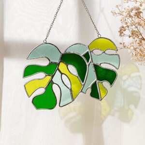 May include: Stained glass Monstera leaf suncatcher with a chain for hanging. The design features shades of green, yellow, and light blue. The glass is outlined with a dark metal. A decorative piece for a window or wall.