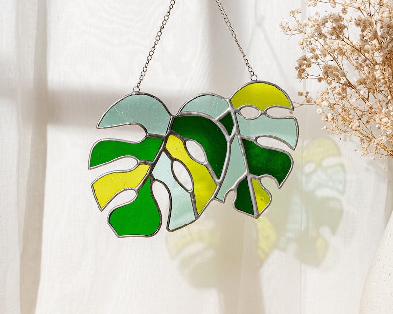 May include: Stained glass Monstera leaf suncatcher with a chain for hanging. The design features shades of green, yellow, and light blue. The glass is outlined with a dark metal. A decorative piece for a window or wall.
