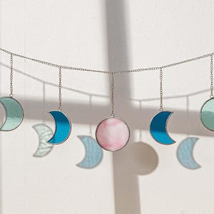 Puede incluir: Una guirnalda decorativa de fases lunares con lunas crecientes y una luna llena. Las lunas son de vidrieras en tonos de azul, verde y rosa, suspendidas de una cadena plateada. Para la decoraci&oacute;n de paredes.