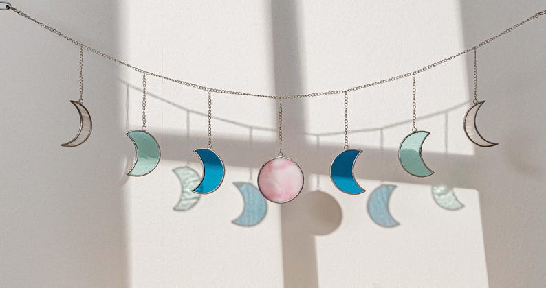 Puede incluir: Una guirnalda decorativa de fases lunares con lunas crecientes y una luna llena. Las lunas son de vidrieras en tonos de azul, verde y rosa, suspendidas de una cadena plateada. Para la decoraci&oacute;n de paredes.
