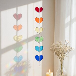Op de afbeelding: Een regenboogkleurige hartvormige zonnevanger die aan een ketting hangt. De zonnevanger heeft zeven harten in rood, oranje, geel, groen, blauw en indigo. De glazen harten zijn verbonden door een zilveren ketting en een kleine kristal aan de onderkant.