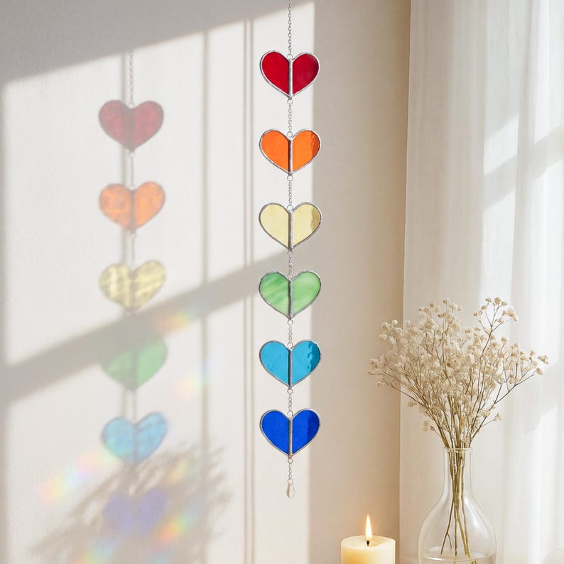 Op de afbeelding: Een regenboogkleurige hartvormige zonnevanger die aan een ketting hangt. De zonnevanger heeft zeven harten in rood, oranje, geel, groen, blauw en indigo. De glazen harten zijn verbonden door een zilveren ketting en een kleine kristal aan de onderkant.