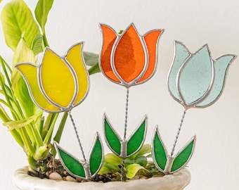 Könnte beinhalten: Drei Gartenstecker in Form von Tulpen aus farbigem Glas. Die Tulpen sind gelb, orange und hellblau, mit grünen Blättern. Die Stiele sind aus silbernem Draht. Sie stehen in einem weißen Topf mit einer grünen Pflanze.