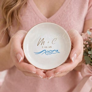 Puede incluir: Un pequeño plato redondo de cerámica blanca con borde dorado. El plato presenta las iniciales "M + C" en escritura marrón, la fecha "7.22.26" e ilustraciones de olas azules. El plato se sostiene con ambas manos.