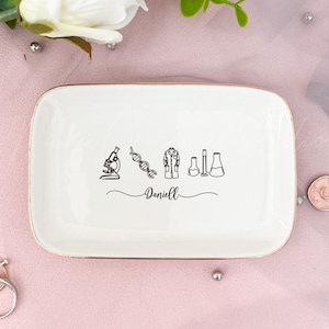 Può includere: Un vassoio rettangolare bianco con bordo dorato. Il piatto presenta disegni a tratto nero di icone legate alla scienza e il nome "Daniell". Sono raffigurati un microscopio, una catena di DNA, un camice da laboratorio e becher.