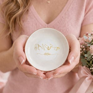 May include: A small, round, white ceramic dish with a gold rim and gold line art of nail tools. The dish has the word "Ritika" in gold script. The dish is held in front of a pink top and a bouquet of pink roses.