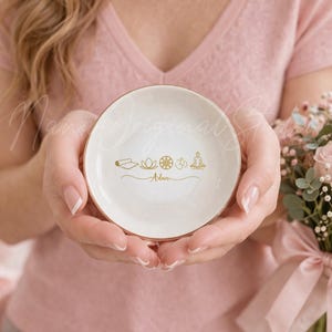May include: A small, round, white ceramic dish with a gold rim. The dish features gold line art of yoga-related symbols and the name "Adam" in script. The dish is held in front of a pink background, with a bouquet of flowers to the right.