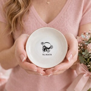 May include: A small, round, white ceramic trinket dish with a metallic gold rim. The center features a black line-art illustration of a human skull surrounded by roses, with the text "TIL DEATH" printed in black capital letters below it.