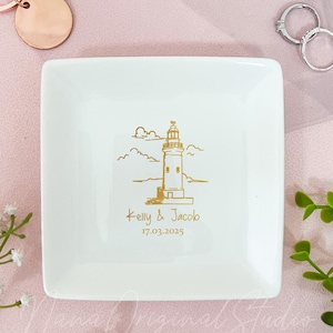 May include: A white square ceramic ring dish with a gold lighthouse design, the names "Kelly & Jacob" and the date "17.03.2025". The dish is surrounded by a rose gold keychain, rings, and greenery.