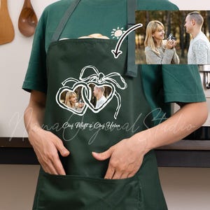 Puede incluir: Un delantal verde oscuro con un bolsillo frontal grande y un gráfico de líneas blancas de dos corazones entrelazados unidos por un lazo. Dentro de los corazones hay dos inserciones fotográficas circulares que muestran a una pareja. El texto "Chef Matt & Chef Helen" está impreso debajo.