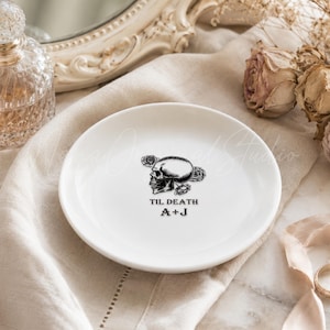 May include: A white ceramic trinket dish featuring a black graphic of a skull surrounded by roses. The text "TIL DEATH" is printed above the initials "A+J" in a black serif font centered on the surface of the round dish.