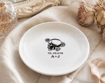 May include: A white ceramic trinket dish featuring a black graphic of a skull surrounded by roses. The text "TIL DEATH" is printed above the initials "A+J" in a black serif font centered on the surface of the round dish.