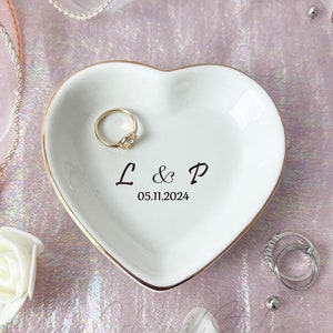 May include: White heart-shaped ceramic ring dish with a gold trim. The dish has the initials "L & P" and the date "05.11.2024" written on it. A gold ring is sitting in the dish.