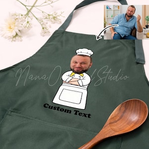 May include: Dark green apron with a cartoon chef graphic and the text "Custom Text". The chef graphic features a person's face, a white chef's hat, and a white chef's coat with a yellow bow tie. A wooden spoon rests on the apron.