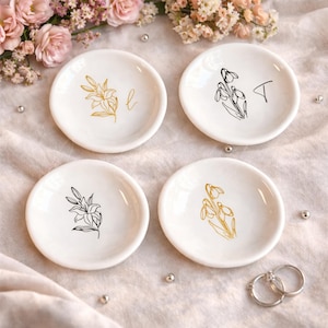 May include: Four small, white ceramic ring dishes. Each dish features a floral design in gold or black, with a single initial on two of the dishes. The dishes are arranged on a soft, light-coloured surface, with flowers in the background.