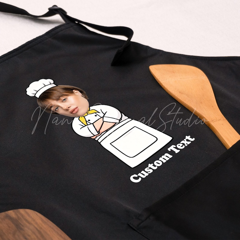 May include: Black apron featuring a cartoon chef with a custom text area. The chef illustration includes a chef's hat and a white apron. A wooden spoon rests on the apron. The text "Custom Text" is printed in white.