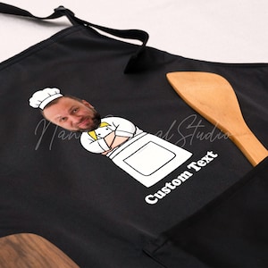 May include: Black apron featuring a cartoon chef with a person's face, a white chef's hat, and the words "Custom Text" in white. A wooden spoon rests on the apron. The apron is designed for kitchen use.
