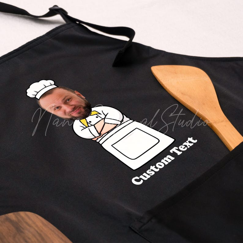 May include: Black apron featuring a cartoon chef with a person's face, a white chef's hat, and the words "Custom Text" in white. A wooden spoon rests on the apron. The apron is designed for kitchen use.