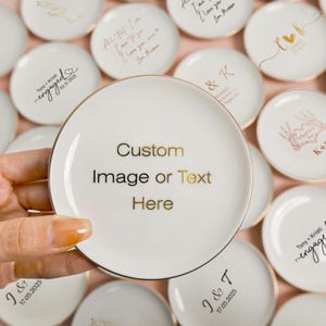 May include: A round white ceramic trinket dish with a gold-colored rim. The center of the dish features the gold-colored text "Custom Image or Text Here" printed in a clean, sans-serif font. The dish is surrounded by other similar dishes.