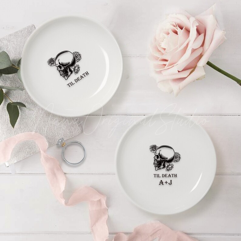 May include: Two white ceramic ring dishes featuring a black graphic of a skull surrounded by roses. One dish displays the text TIL DEATH, while the other features TIL DEATH and the initials A+J in black lettering.