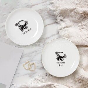 May include: Two white ceramic plates featuring a black graphic of a skull surrounded by roses. One plate displays the text TIL DEATH, while the other features the text TIL DEATH and the initials A+J in black lettering.