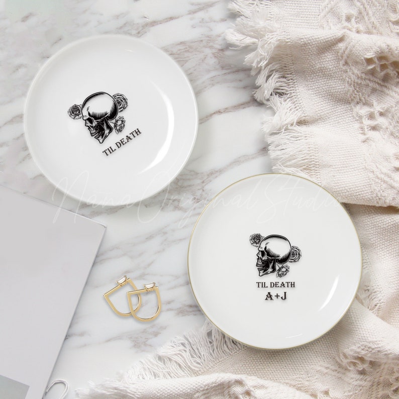 May include: Two white ceramic plates featuring a black graphic of a skull surrounded by roses. One plate displays the text TIL DEATH, while the other features the text TIL DEATH and the initials A+J in black lettering.