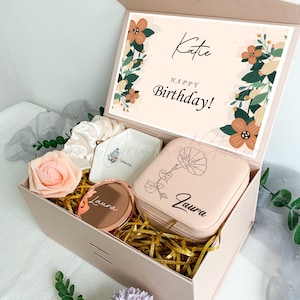 Puede incluir: Una caja de regalo rosa con un dise&ntilde;o floral y el texto "Katie Feliz cumplea&ntilde;os!" en la tapa. La caja contiene una peque&ntilde;a caja de joyer&iacute;a rosa con un dise&ntilde;o floral y el nombre "Laura" grabado en ella, un peque&ntilde;o plato blanco con el nombre "Laura" en &eacute;l, un peque&ntilde;o espejo redondo con el nombre "Laura" en &eacute;l y una diadema rosa.