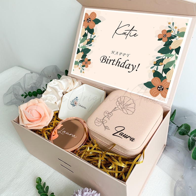 Puede incluir: Una caja de regalo rosa con un dise&ntilde;o floral y el texto "Katie Feliz cumplea&ntilde;os!" en la tapa. La caja contiene una peque&ntilde;a caja de joyer&iacute;a rosa con un dise&ntilde;o floral y el nombre "Laura" grabado en ella, un peque&ntilde;o plato blanco con el nombre "Laura" en &eacute;l, un peque&ntilde;o espejo redondo con el nombre "Laura" en &eacute;l y una diadema rosa.