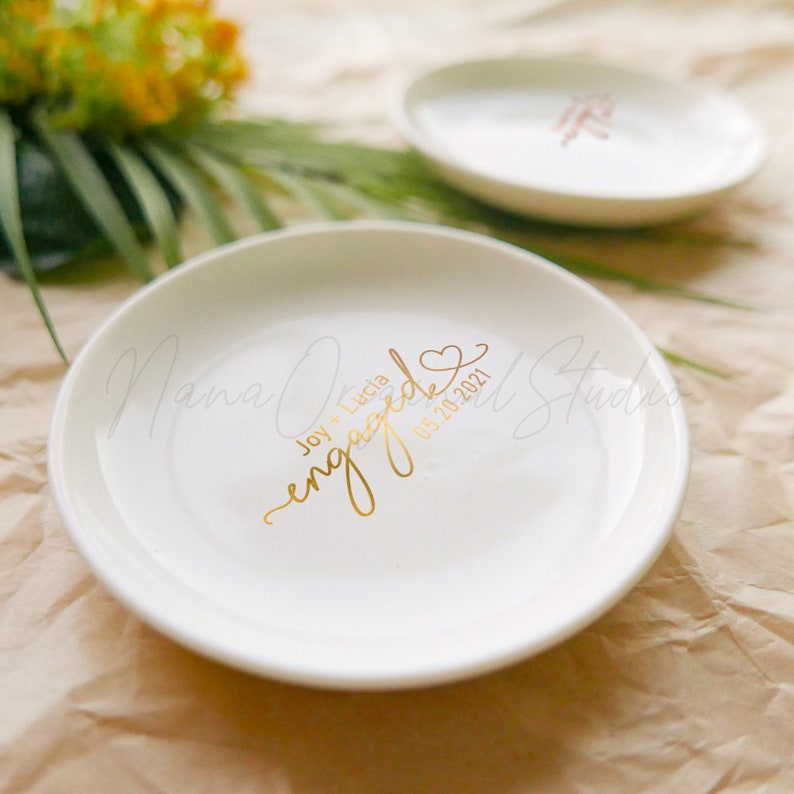 May include: Two white ceramic ring dishes with gold lettering. The front dish says "Joy + Lucia engaged 05.20.2021" in gold script. The second dish is visible in the background. The dishes are on a brown paper surface.