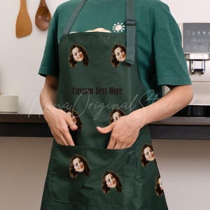 May include: Dark green apron featuring multiple smiling face graphics. The apron has a large front pocket and an adjustable neck strap. The text "Custom Text Here" is printed on the apron. A small sun graphic is on the chest.