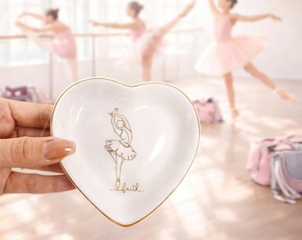 May include: A heart-shaped white ceramic trinket dish with a gold rim and a gold ballerina illustration. The word "faith" is written in gold script below the ballerina. The background shows a ballet studio with dancers in pink tutus.