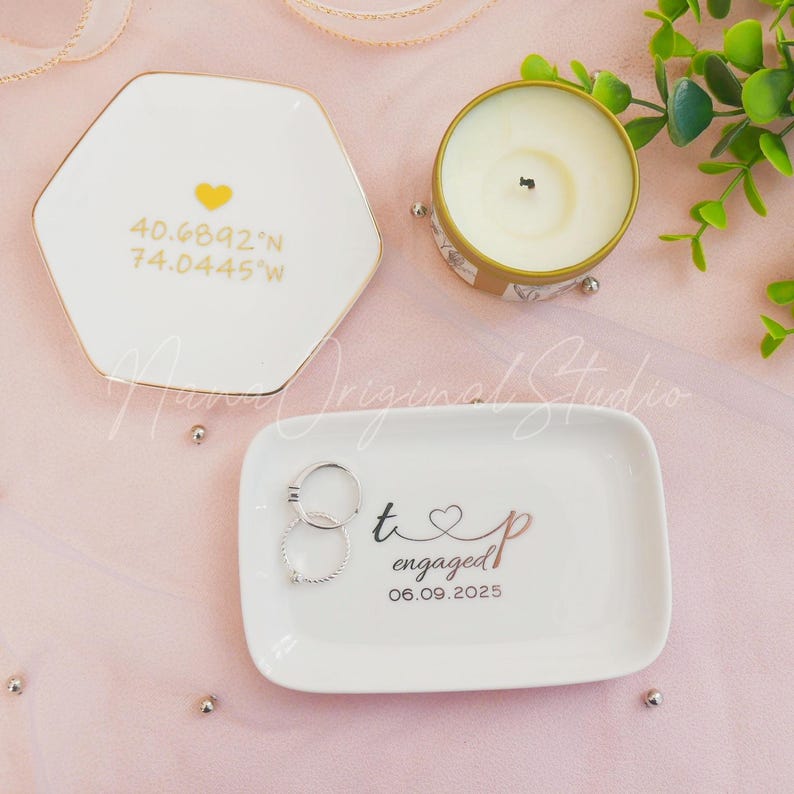 May include: White ceramic ring dishes with gold trim, one hexagonal with coordinates and a heart, the other rectangular with rings and the word "engaged" and a date. A small candle in a gold tin is also present.